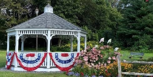 The white Town of Charlton gazebo, draped in red, white and blue bunting, with pink hydrangeas beside it